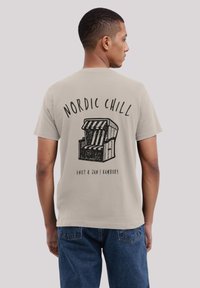 Mann trägt ein beiges T-Shirt mit dem Aufdruck "NORDIC CHILL" und einer Grafik eines gestreiften Strandstuhls, kombiniert mit blauen Jeans.