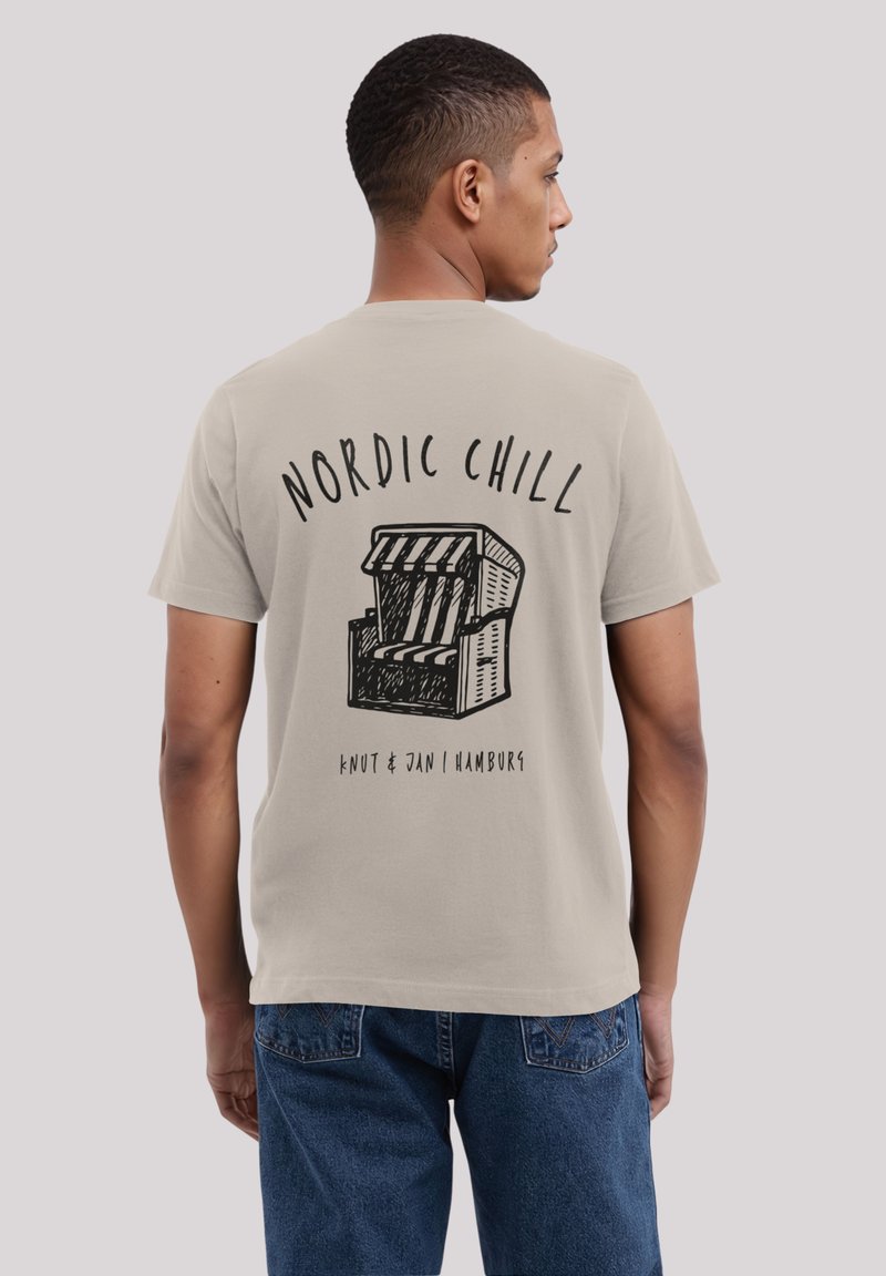 Mann trägt ein beiges T-Shirt mit dem Aufdruck "NORDIC CHILL" und einer Grafik eines gestreiften Strandstuhls, kombiniert mit blauen Jeans.