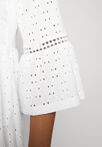 Robe blanche brodée avec motif ajouré, dotée de manches évasées, d'une finition texturée et d'une taille cintrée avec des détails découpés.