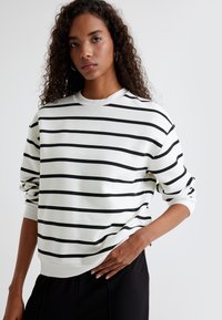 Sudadera blanca con rayas horizontales negras, cuello redondo, puños de canalé, corte relajado, textura de tela suave, combinada con pantalones negros.