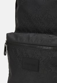 Zaino in tessuto nero con motivo a logo ripetuto, tasca frontale con zipper e un'etichetta logo nera testurizzata nella parte inferiore.