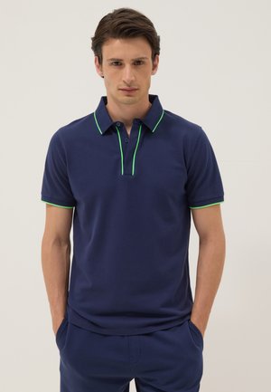 Man staat recht voor zich uit kijkend, draagt een marineblauw poloshirt met neongroene bies aan kraag, knoopsluiting en mouwen, handen in de zakken.