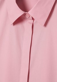 Jasnoróżowa koszula z wywiniętym kołnierzykiem, jedynym zapięciem na guzik i gładką fakturą. Posiada pionową szew wzdłuż przodu.
