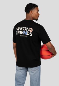 Černé bavlněné tričko s vícebarevným grafikou "WRONG FRIENDS DESIGN HOUSE" na zadní straně. Model drží basketbalový míč a má na sobě světle modré džíny.