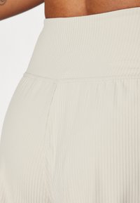 Beige ribbet stoff med bredt belte og subtile folder. Glatt tekstur med en lett glød, designet for komfort og fleksibilitet.