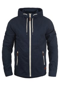 Veste bleu marine en coton avec une fermeture éclair. Comprend deux poches avant, une capuche avec cordon de serrage et des accents en cuir.