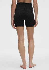 Person, der står barfodet og vender væk, iført høj talje sorte shorts med små prikker på en ensfarvet baggrund.