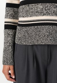 Pruhovaný pletený sveter v čiernej, bielej a béžovej farbe; má štruktúrovaný povrch a široké manžety. Skombinovaný s elegantnými šedými nohavicami.