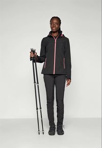 Femme portant une veste et un pantalon de randonnée noirs et roses, tenant deux bâtons de trekking noirs, se tenant devant un fond clair.