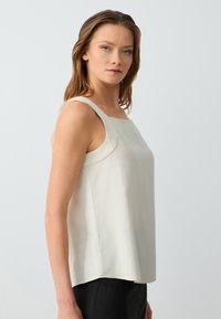 Donna con capelli castano chiaro, che indossa un top senza maniche color avorio e pantaloni neri, in piedi di lato contro uno sfondo chiaro e semplice.