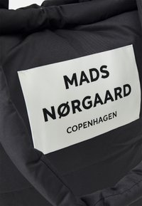 Zwarte tas met een rechthoekig wit label waarop "MADS NØRGAARD COPENHAGEN" in vetgedrukte zwarte tekst staat. Gestructureerde stof en gevoerde handvatten.