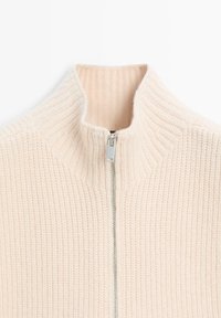 Krämfärgad, ribbad stickad tröja med hög krage och en silverdragkedja delvis dragit upp framtill i mitten.