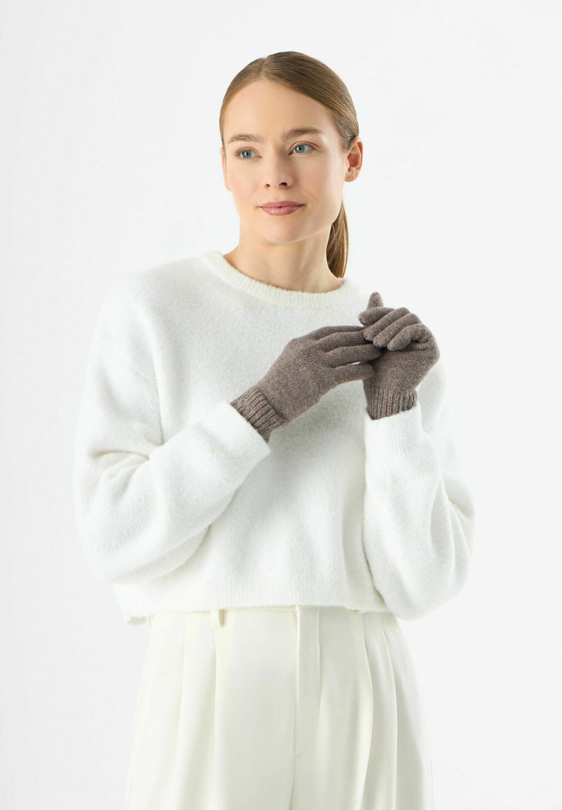 Gants tricotés gris avec des poignets côtelés, portés par un modèle. Les gants présentent un design texturé, associés à un pull en polaire blanc.