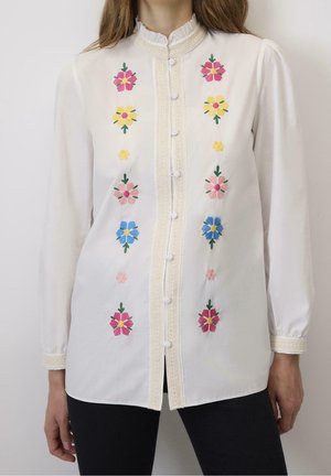 Hvid bluse med lange ærmer med lodrette rækker af farverige broderede blomster og stofbetrukne knapper ned langs midten.