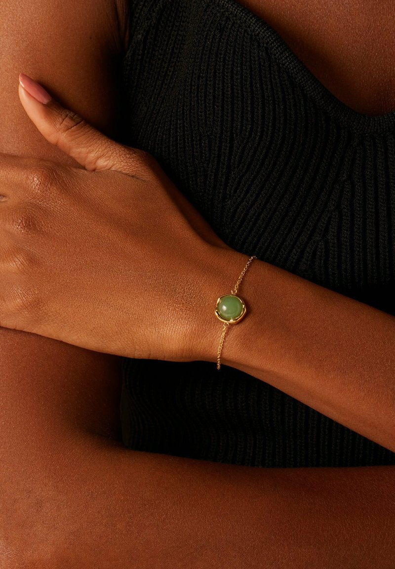 Goldene Gliederarmband mit einem oktagonalen grünen Stein als Mittelpunkt, getragen an einem dunkelhäutigen Handgelenk, vor einem strukturierten schwarzen Hintergrund.