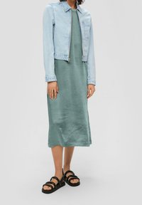 Hellblaues Denim-Jacket über einem grünen Satin-Midikleid; Modell trägt schwarze Riemchensandalen. Glatte Texturen, Kontrast in den Stoffoberflächen.