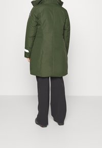 Parka verde acolchada con un cuello alto, detalles en negro y franjas reflectantes blancas en las mangas, combinada con pantalones anchos negros.