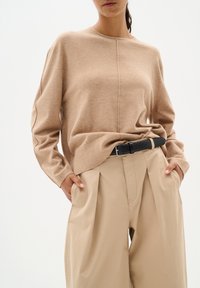 Beżowy sweter wełniany z okrągłym dekoltem, luźny krój i widocznymi szwami, w połączeniu z beżowymi spodniami o wysokim stanie i czarnym paskiem.