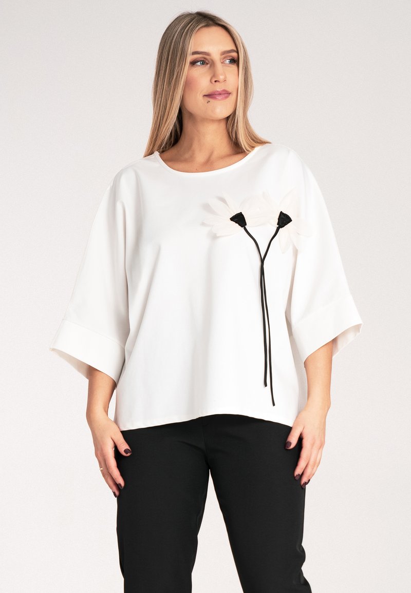 Biały top z okrągłym dekoltem i luźnymi rękawami, ozdobiony czarnymi aplikacjami w kwiaty oraz łodygami, zestawiony z czarnymi spodniami. Gładka faktura materiału.