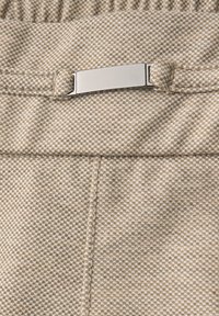 Béžová a bílá tkanina s kostkovaným vzorem houndstooth, s obdélníkovou stříbrnou kovovou sponou připevněnou k odpovídajícímu poutku na pásek.