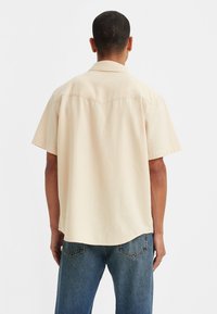 Short-sleeve, cream-colored shirt with a relaxed fit, collar, and curved hem, paired with light blue denim jeans, viewed from the back.