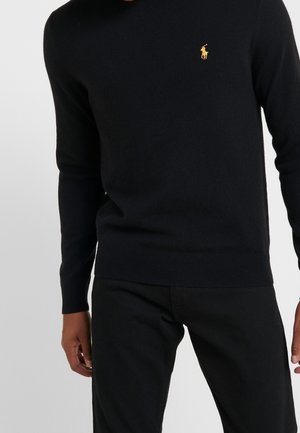 Personne portant un pull ajusté à manches longues noir avec un petit logo brodé représentant un cheval jaune sur la poitrine, associé à un pantalon noir.