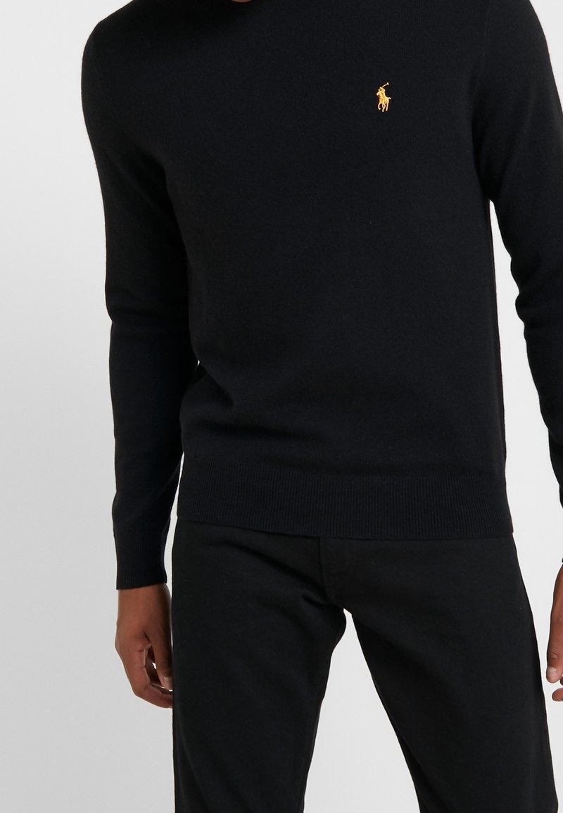 Personne portant un pull ajusté à manches longues noir avec un petit logo brodé représentant un cheval jaune sur la poitrine, associé à un pantalon noir.
