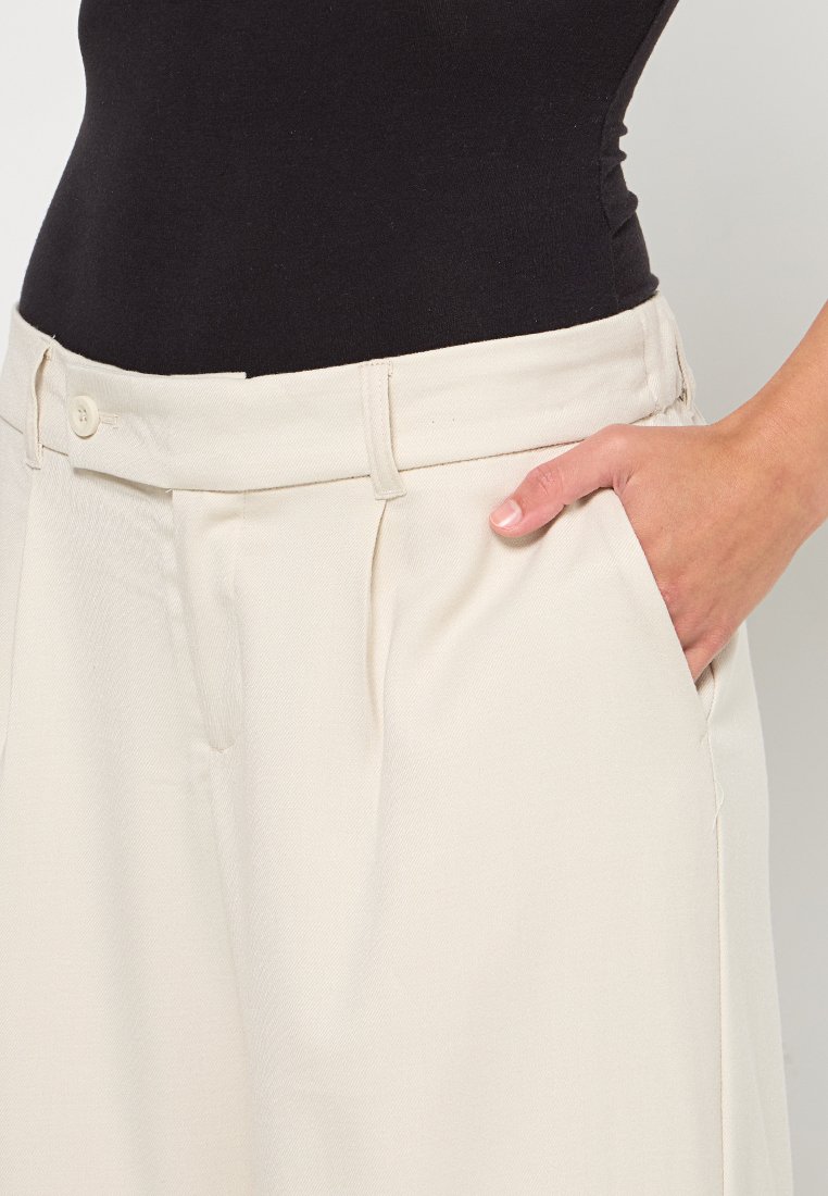 Beige weite Hosen mit glatter Textur, ausgestattet mit Seitentaschen und einem Knopfverschluss am Bund. Taillierte Passform an der Taille.