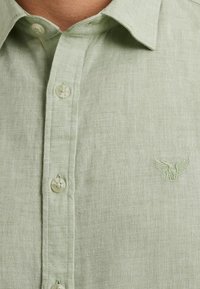 Lichtgroen linnen overhemd met een button-down kraag, voorzien van doorzichtige knopen en een subtiel geborduurd arendlogo op de linkerborst.
