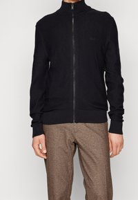 Stickad svart zip-up jacka med hög krage, med ribbade ärmslut och nederkant. Diskret logotypdetalj på bröstet. Slen textur.