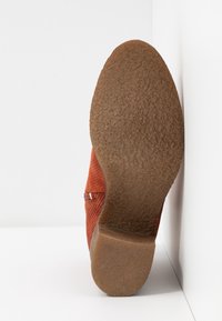 Semelle d'une chaussure marron avec semelle extérieure en caoutchouc texturé et petit talon, placée verticalement contre une surface blanche en angle.