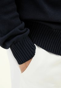 Marinblå stickad tröja med ribbade muddar. Bomullsblandad väv, slät textur. Vita byxor synliga i bakgrunden, med en hand i fickan.