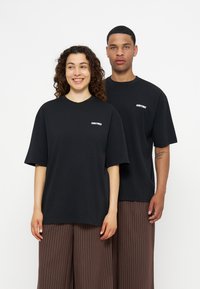 Δύο μοντέλα φορούν μαύρα φαρδιά T-shirts με λευκό λογότυπο και καφέ ριγέ παντελόνες με φαρδιά πόδια, σε απλό λευκό φόντο.