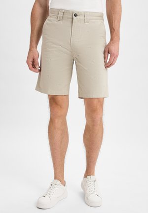 Man in beige shorts met kleine vis borduursels, witte sneakers en een wit shirt, staande tegen een effen achtergrond.