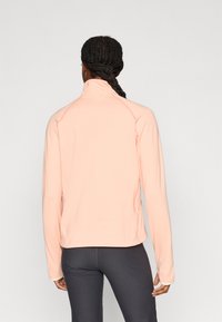 Ανοιχτό ροζ ζακέτα με φερμουάρ, μακριά μανίκια, υφή υφάσματος και ψηλό γιακά. Φορεμένη με σκουρόχρωμες κολάν, που δείχνει εφαρμοστή γραμμή.