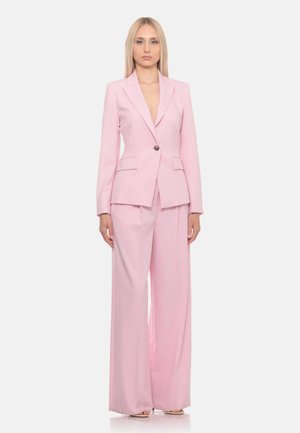 Donna bionda in piedi, indossa un blazer rosa chiaro su misura con un bottone e pantaloni a gamba larga abbinati su uno sfondo uniforme.
