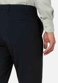 Marineblauwe dressbroek met een gladde textuur, slim fit en een enkele achterzak. Een lichtgekleurde blouse is gedeeltelijk zichtbaar.