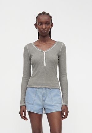 Žena s vrkočmi, oblečená v sivé dlhé henley tričko a svetlomodré rifľové šortky, stojaca pred jednoliatym bielym pozadím.