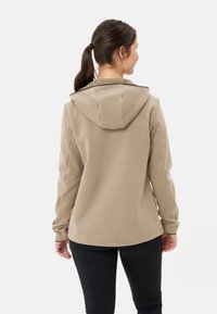 Beige Kapuzenjacke aus strukturiertem Stoff, mit langen Ärmeln und kontrastierenden schwarzen Akzenten, kombiniert mit schwarzen engen Hosen. Klare Designlinien und Struktur.
