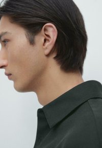 Dunkelgrünes Poloshirt aus strukturiertem Stoff mit einem umgeschlagenen Kragen und Seitenansicht des Kopfes eines männlichen Models. Glattes Haar und reine Haut.