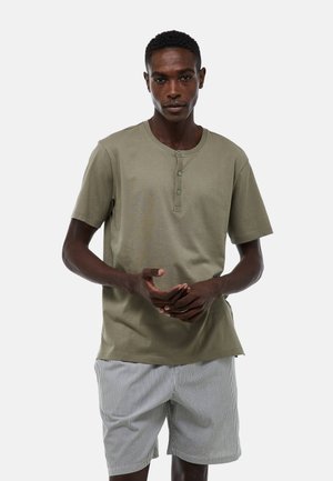 Homme portant un t-shirt henley manches courtes vert olive et un short blanc avec de fines rayures noires, debout devant un fond clair uni.