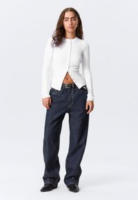 Cardigan blanc à manches longues et côtelé avec fermeture par boutons, associé à un jean bleu foncé à jambes larges et des bottes noires à bout pointu.