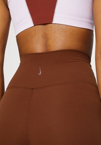 Brown high-waisted leggings with a smooth texture, featuring a small gray Nike logo near the waistband and precise stitching details.