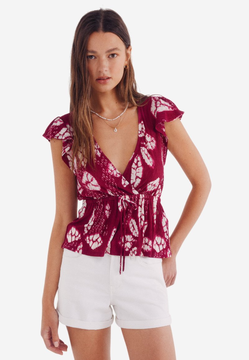Maroone Bluse mit Blumenmuster, Rüschensleeves, tailliertem Detail und tiefem V-Ausschnitt. Kombiniert mit weißen hochgekrempelten Shorts.