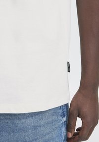 White cotton t-shirt with a straight cut, featuring a small black tag on the side. Paired with light blue denim jeans.