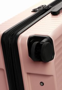 Světle růžový tvrdý kufr s černými zipy a akcenty, s drážkovaným povrchem a mechanismem dvojitých koleček.