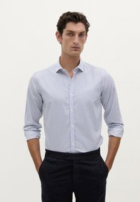 Cămașă cu nasturi de culoare albastră deschisă, cu dungi verticale albe. Are un guler clasic și mâneci lungi, asortată cu pantaloni închisi la culoare.