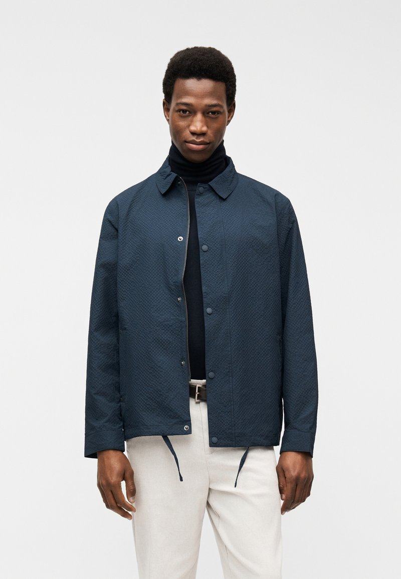 Άνδρας που φοράει υφασμάτινο ναυτικό μπουφάν πάνω από μαύρο ζιβάγκο και ανοιχτό μπεζ παντελόνι με καφέ ζώνη, στέκεται μπροστά σε απλό φόντο.