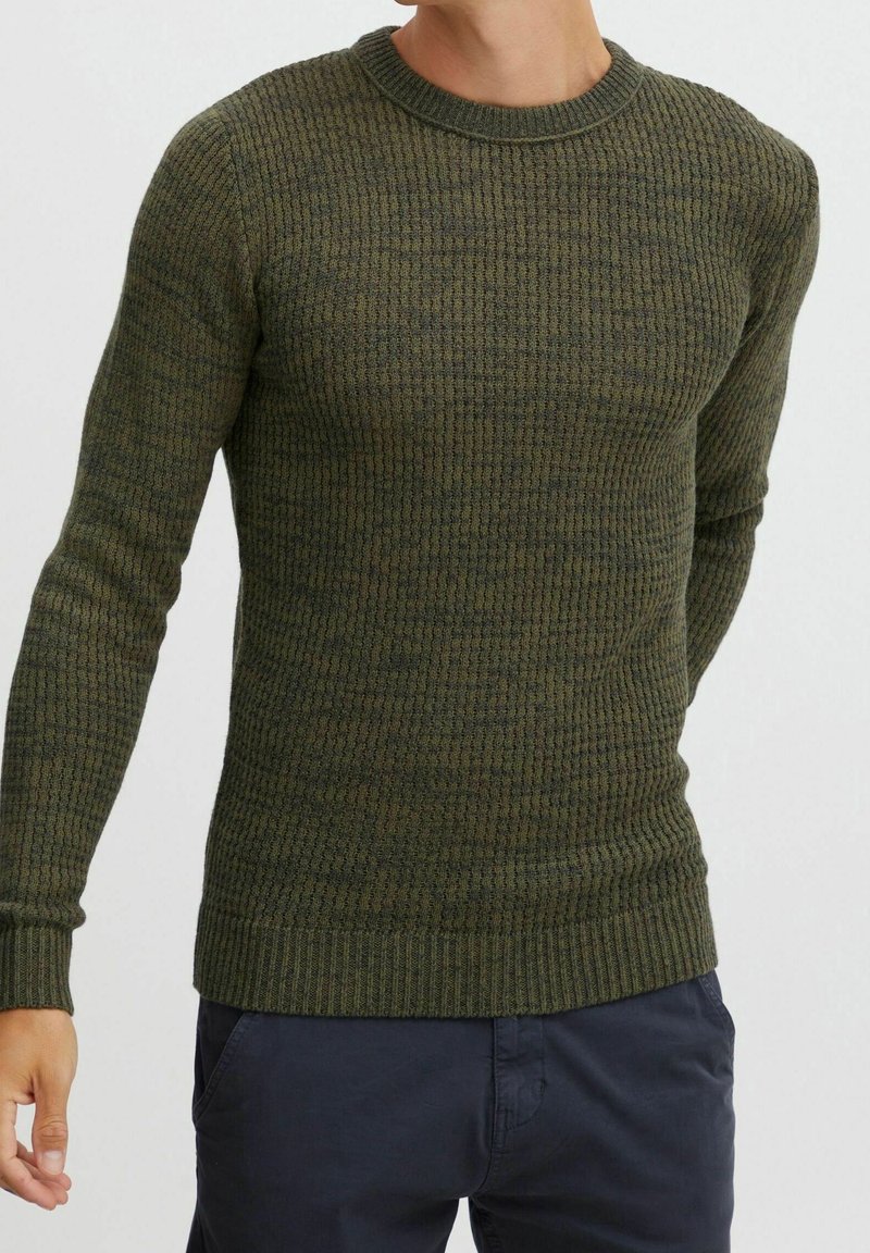 Homme portant un pull ajusté à manches longues en maille texturée vert olive avec un pantalon bleu foncé sur un fond uni.