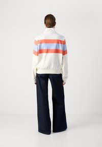Hvit hettejakke med oransje og lyseblå horisontale striper, kombinert med vide mørke denimbukser. Jevn tekstur, avslappet passform.
